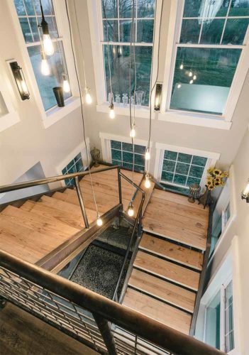 industrial farmhouse style custom new home in St. Charles, Iowa designed and built by Silent Rivers features a grand staircase made of steel channel and bridge plank lumber and hay wheel chandelier custom light fixture with Edison style LED hanging lights