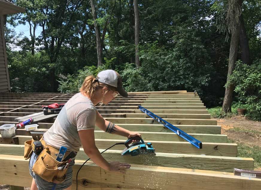 Jenna Frideres, artisan of deck designer and builder Silent Rivers of Des Moines, Iowa, planes joists to ensure the new decking installs seamlessly on this sloped yard leading to the woods.
