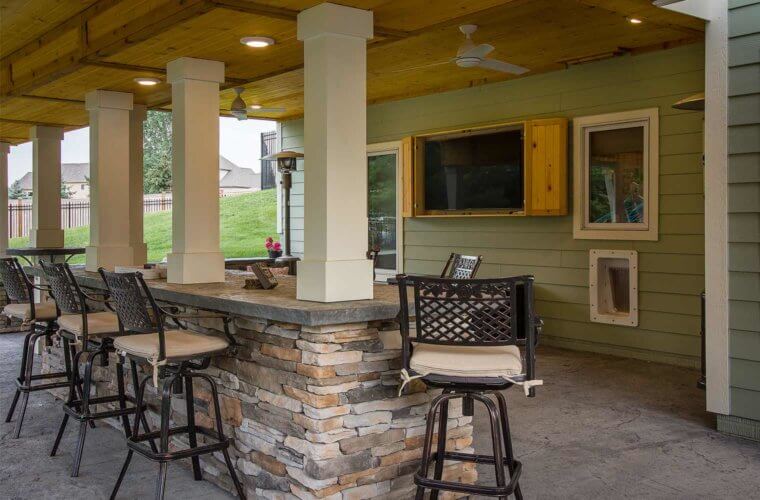 covered patio with curved deck above features outdoor bar area with encased TV and stone accents by builder Silent Rivers Des Moines, Iowa