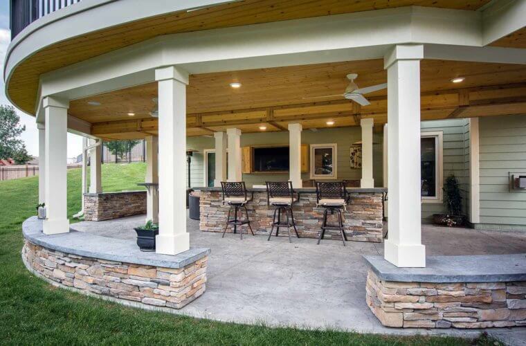 outdoor stone bar with pillars and encased TV in a curved patio underneath a curved deck by builder Silent Rivers, Des Moines, Iowa