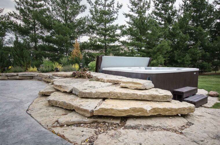 outdoor spa alongside a curved patio surround by slabs of limestone landscaping and nestled among evergreens in outdoor remodel by Silent Rivers, Des Moines, Iowa