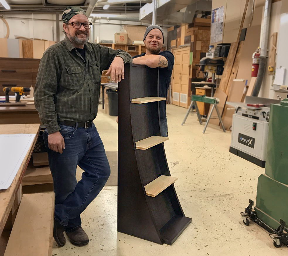 Silent Rivers woodshop artisans Tom Bloxham and Alex Schlepphorst showcase one of their custom designed furniture pieces — a curved bookcase