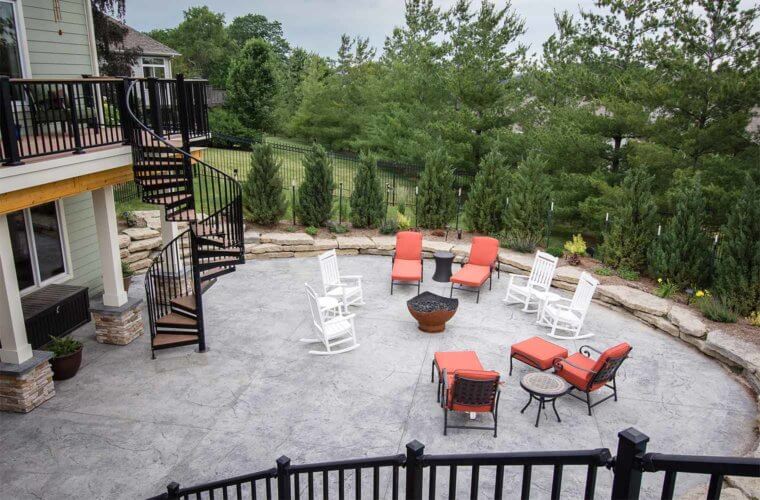 curved metal staircase from upper deck leading to curved patio with fire pot and sitting area surrounded by evergreen trees by builder Silent Rivers Des Moines, Iowa